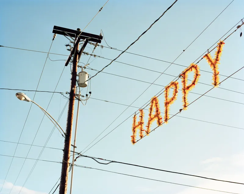 Happy — a lit tinsel sign spelling HAPPY strung between power lines and a utility pole against a clear blue sky. Medium format color film by Alex Hoerner.