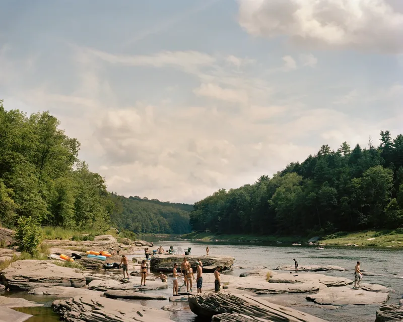 Skinners Falls, NY — summer scene of people swimming and sunning on flat river rocks along the Delaware River, surrounded by dense green forest. Medium format color film by Alex Hoerner.