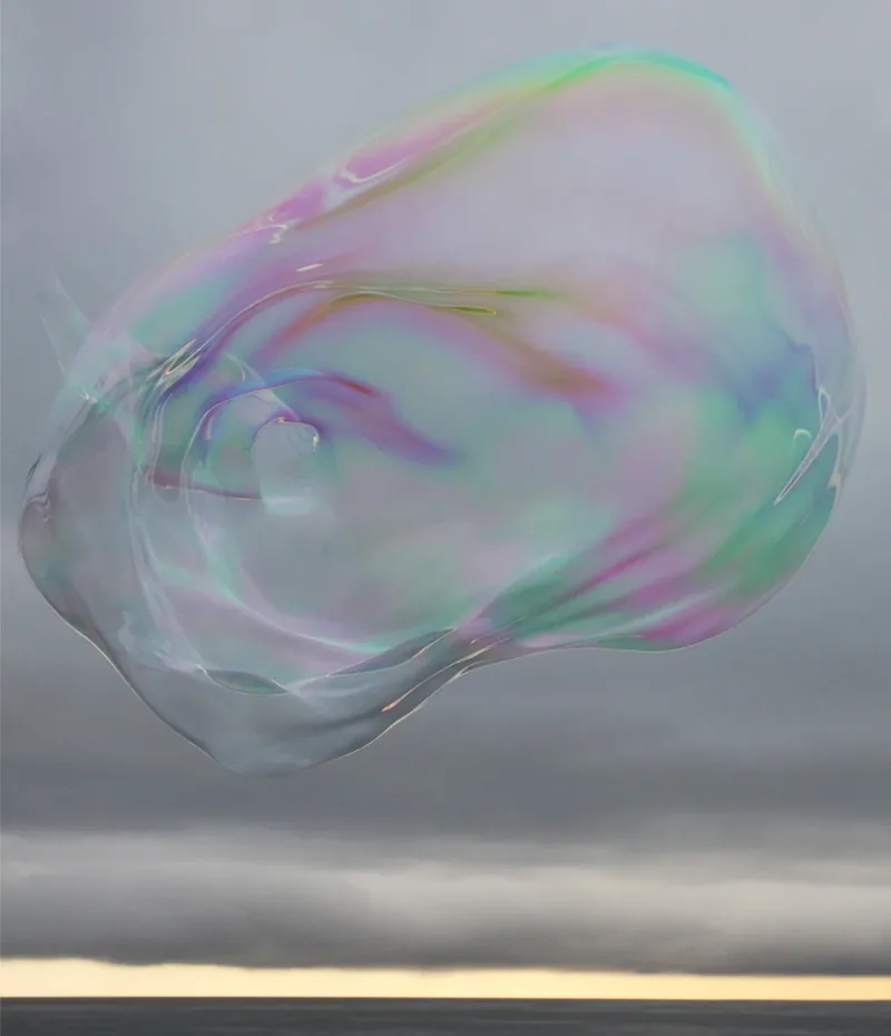 Surface Tension III — close-up of a large iridescent soap bubble with intense pink, green, and lavender swirls of color against a gray sky and ocean horizon. Medium format film by Alex Hoerner.