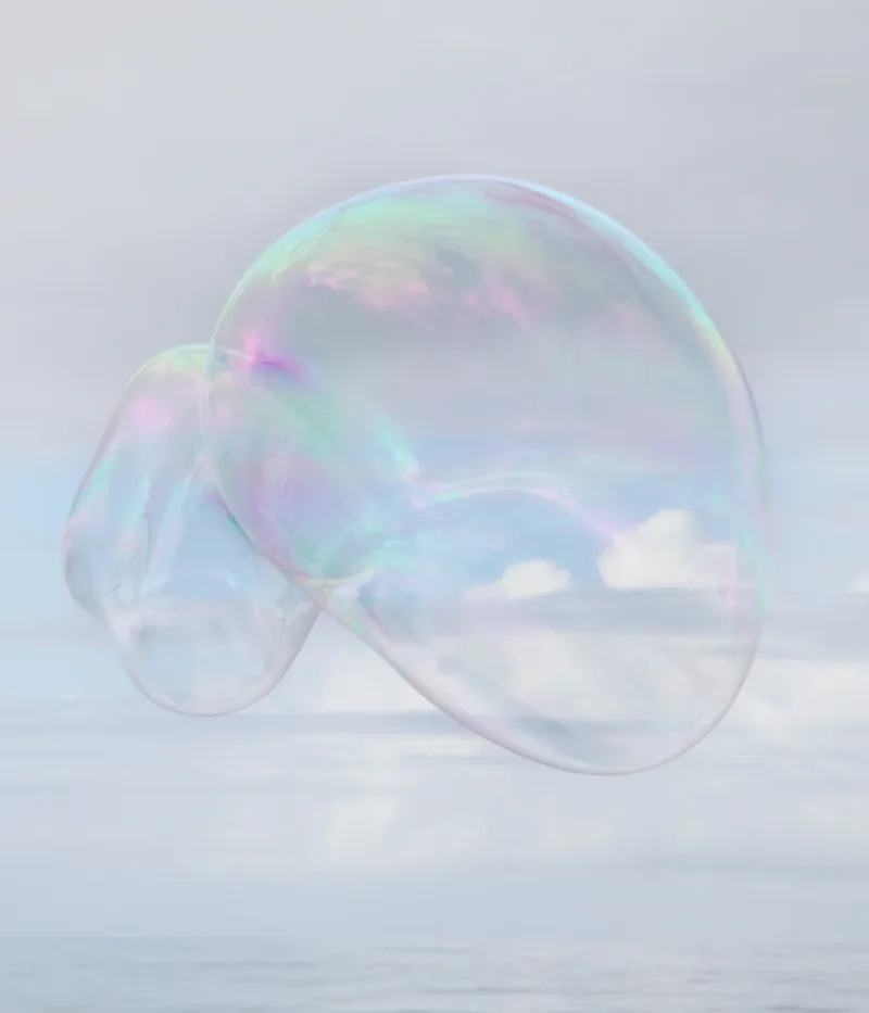 Surface Tension V — two connected iridescent soap bubbles floating against a pale blue-white sky and hazy ocean. Medium format film by Alex Hoerner.